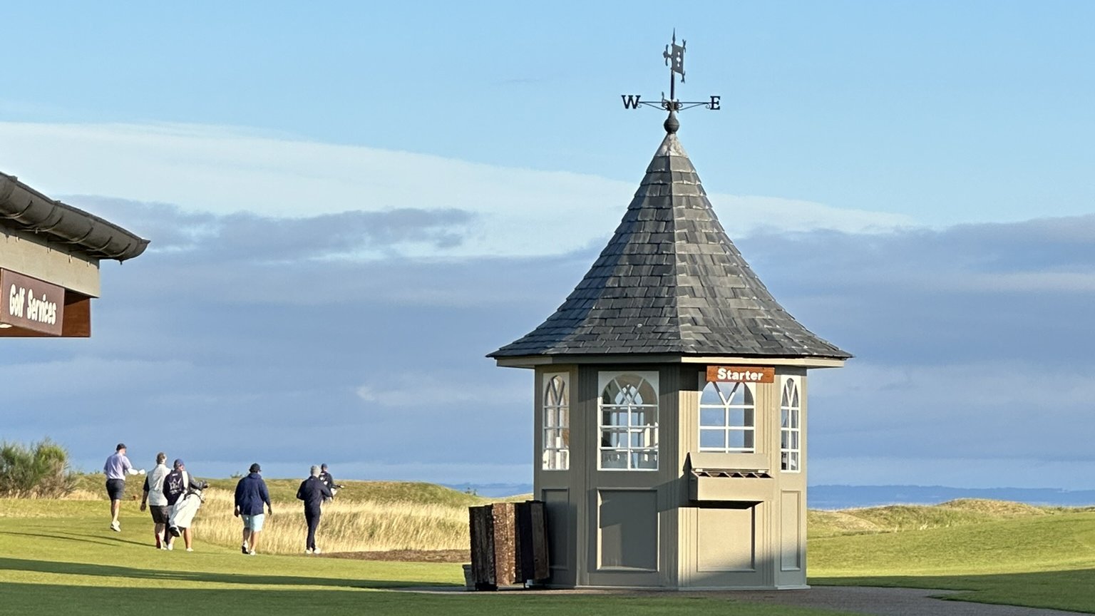 Golfers walking out together onto a green links course past a slate-roofed starter hut with a weathervane under a wide blue-gray sky